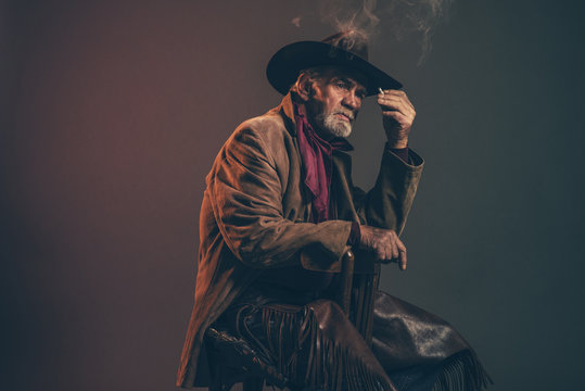 Old rough western cowboy with gray beard and brown hat smoking a