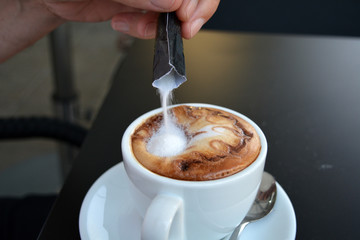 Hand pouring sugar into hot cappuccino