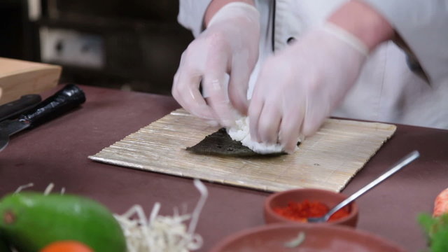Cook making sushi rolls in kitchen, spreading rice over nori