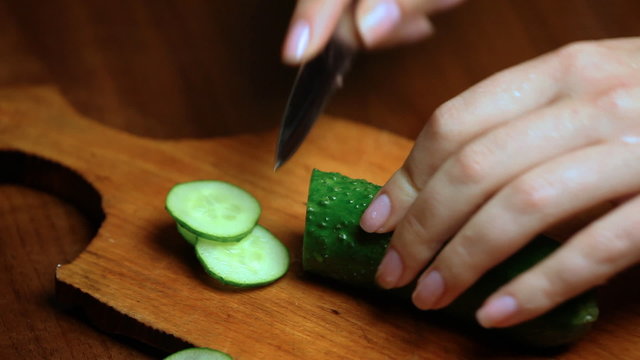 female hand cuts cucumber rings on wooden plate closeup