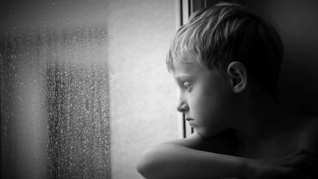 Alone little boy looks raindrops through window glass