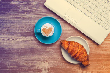 Cup of coffee and croissant near notebook on wooden table.