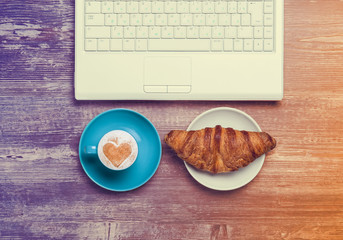 Cup of coffee and croissant near notebook on wooden table.