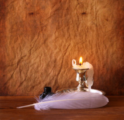 low key image of white Feather, inkwell and burning candle on a 