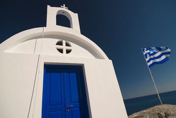 Typical greek scenery with white chapel, blue sea, blue sky and flag.