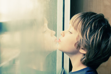 Cute 6 years old boy looking through the window - obrazy, fototapety, plakaty