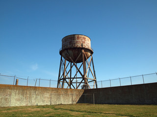 Rusting water tower stands beyond the wall  and bard wire fence