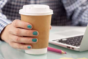 Young woman drinking coffee from disposable cup at her work place while working with a computer 