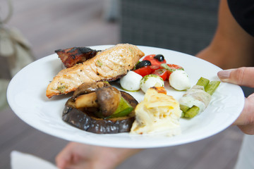 Hand holding a plate with grilled salmon and salad, outside.