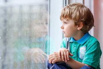 little blond kid boy sitting near window and looking on raindrop