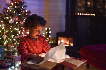 Lovely little girl opens a gift in front of the Christmas tree l