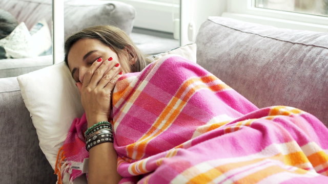 Sick woman coughing, lying on sofa under blanket at home