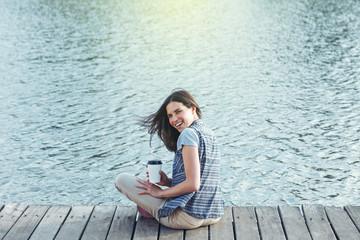 Fototapeta na wymiar beautiful smiling girl with a cup of coffee sitting on a pier