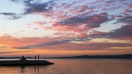 Naklejka na ściany i meble People silhouettes on lake quay at sunset