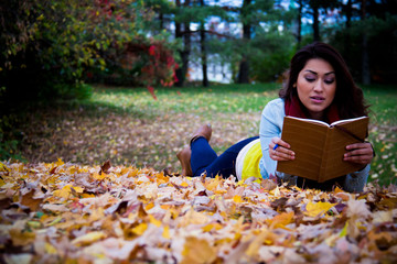 Young woman reading a book lying down on autumn leaves in the fa