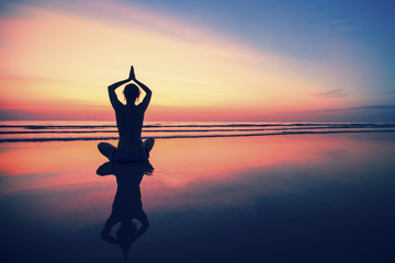 Woman practicing yoga on the sea beach at surrealistic sunset.