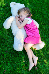 Sad little girl lying on grass with large teddy bear