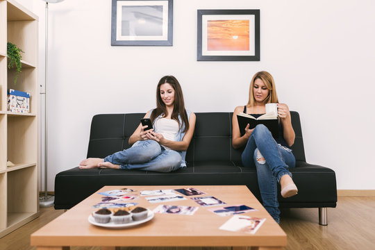 two young women with a smartphone and a book at home