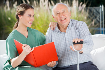 Happy Senior Man Sitting By Female Nurse Holding Book