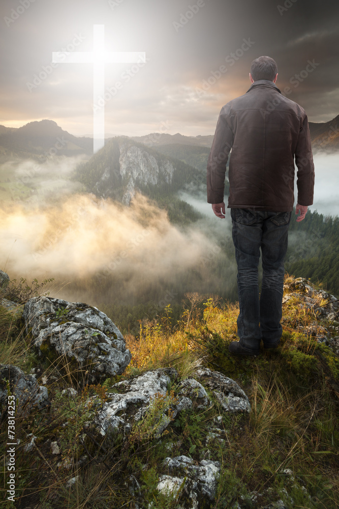 Wall mural Man on top of the mountain looking at Christian Cross at sunrise