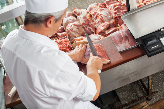 Male Butcher Cutting Meat At Display Cabinet