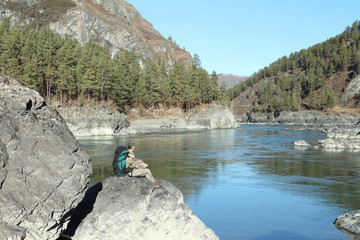 The traveler with a backpack sitting on a stone on the river ban