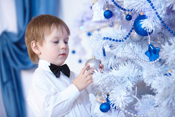 little boy in a white shirt and bow tie decorates a Christmas tr