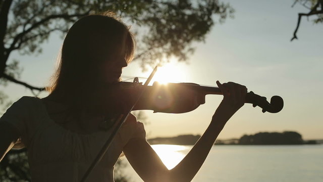 Girl violinist playing the violin at sunset on the lake.