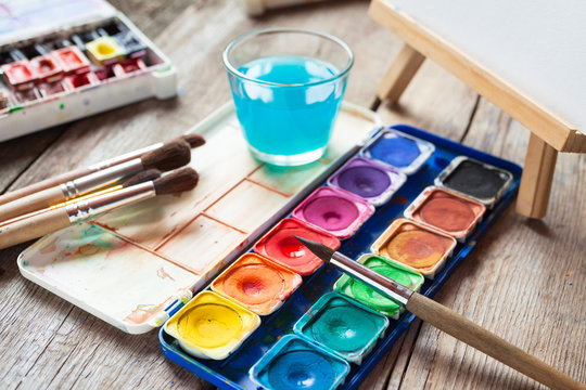Set of watercolor paints, art brushes, glass of water and easel