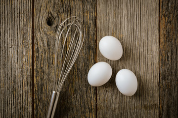 Three Eggs and Whisk or Egg Beater on Rustic Wood Background