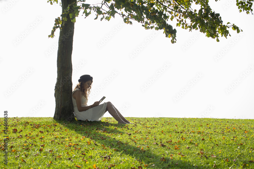 Wall mural attractive young woman reading a tablet under a tree