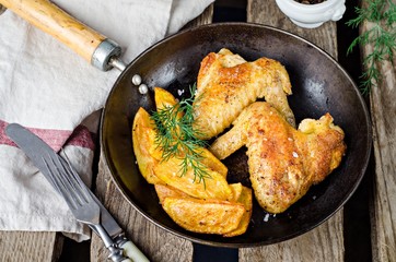 Roasted chicken wings with fried potatoes and ketchup in iron pot on wooden vintage background