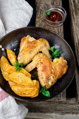 Roasted chicken wings with fried potatoes and ketchup in iron pot on wooden vintage background
