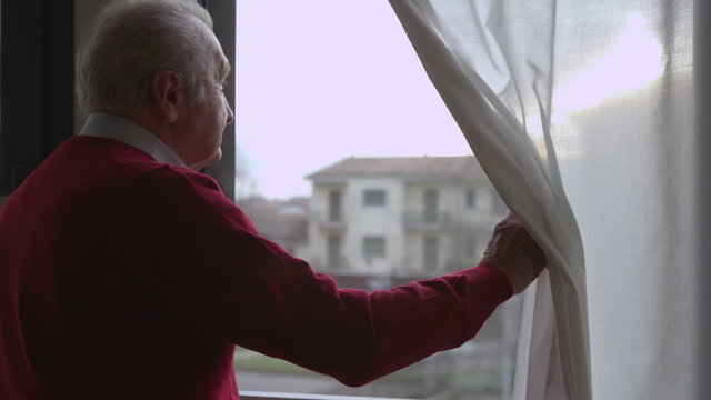 Worried elderly man moves the curtain and looks out the window