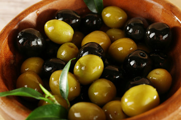 Black and green olives with leaves in bowl on wooden background