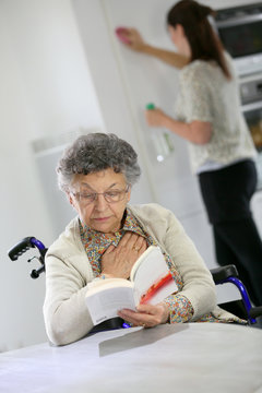 Elderly woman reading book, home helper in background
