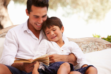 Father And Son Sitting In Garden Reading Book Together