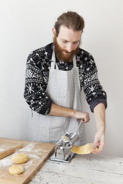 bearded man with apron making fresh pasta using pasta machine
