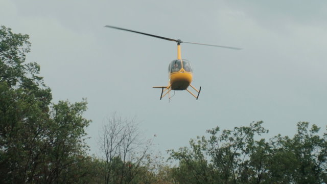 A yellow small manned helicopter hung in the air above trees in the forest. Rescue helicopter