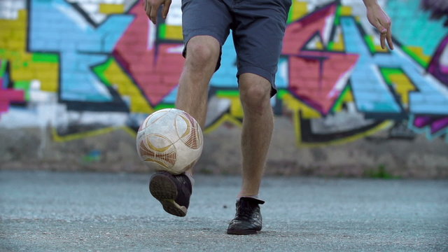 Knee level shot of unrecognizable young sportsman kicking football outdoors, colorful graffiti visible in background