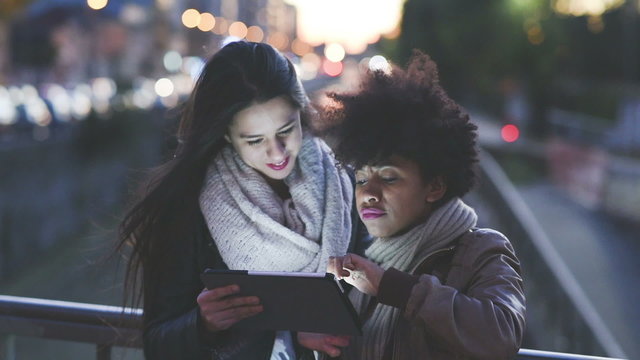 two young beautiful multiethnic girls using tablet by night 