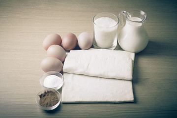 Milk, sugar, cocoa, puff dough, eggs and salt on a wooden board.