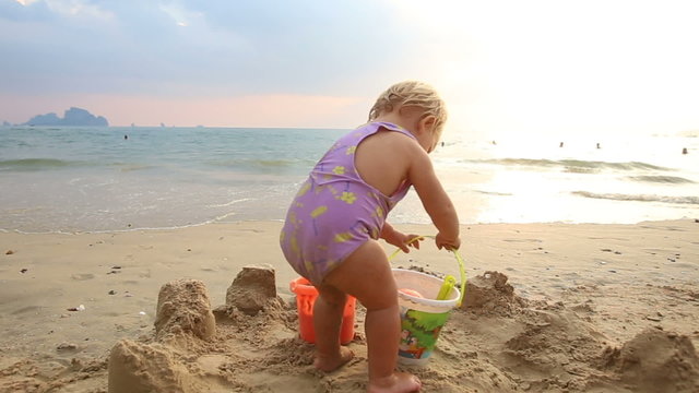 small baby girl in purple swimsuit put the bucket on send and fa