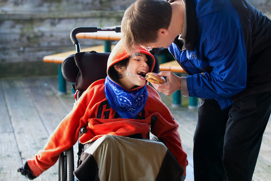 Father feeding disabled son a hamburger in wheelchair. Child has