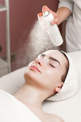 serum mask spraying from can on young woman's face in spa salon