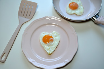 Heart shaped egg on a pink plate with a pink pan and spatula.