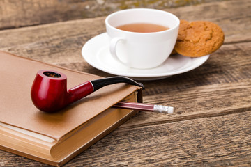 ivory tea cup with sweet cookie, tobacco pipe and book on wooden