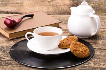 ivory tea cup with sweet cookie, book and tobacco pipe on wooden