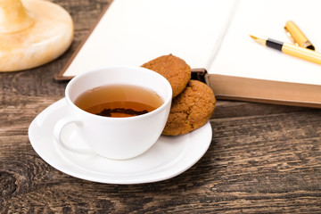 ivory tea cup with sweet cookie, pen and book on wooden palette