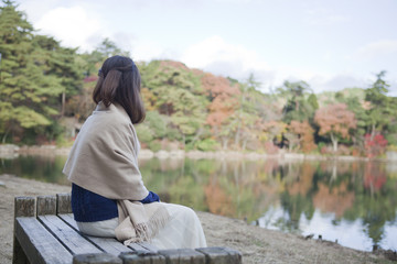Naklejka na ściany i meble 秋の紅葉の公園の池の前のベンチに座る女性